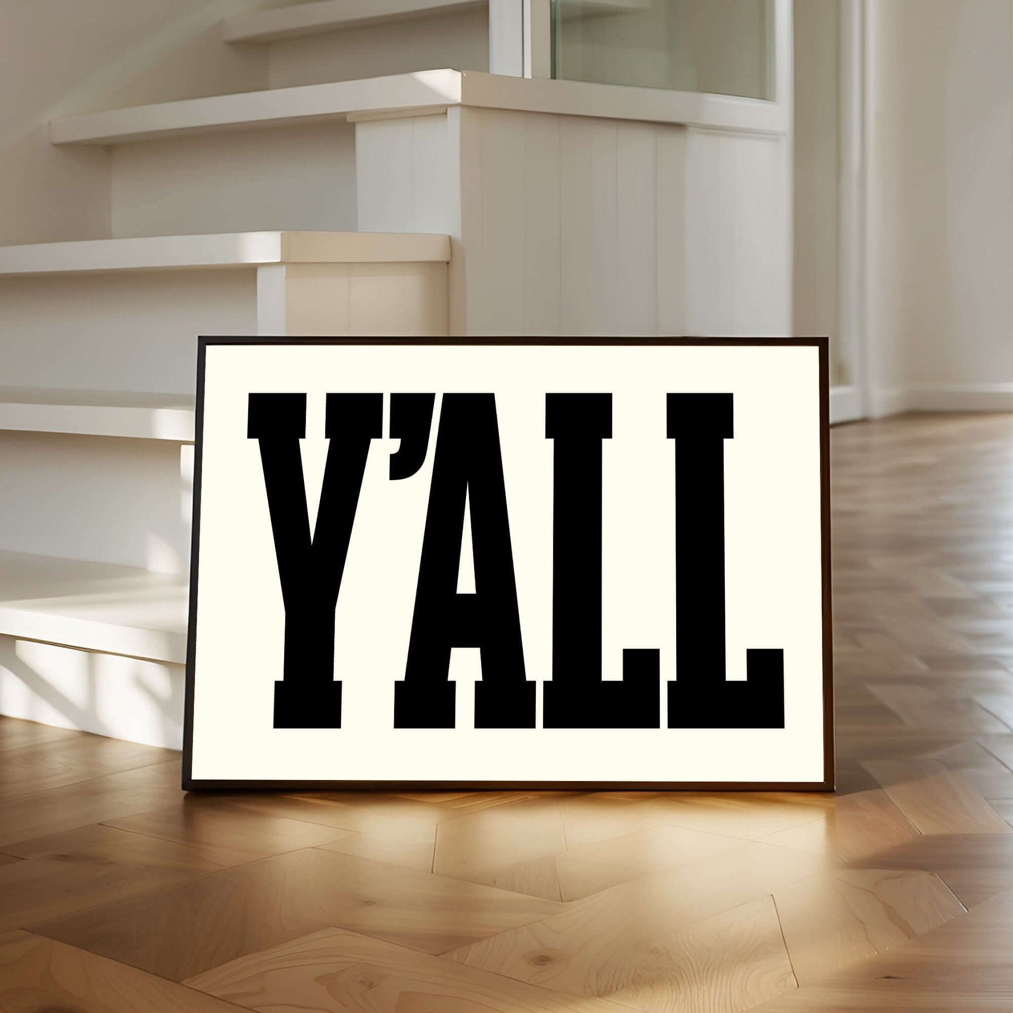 A framed sign with the word "Y'ALL" in bold black letters sits on a wooden floor by white stairs.