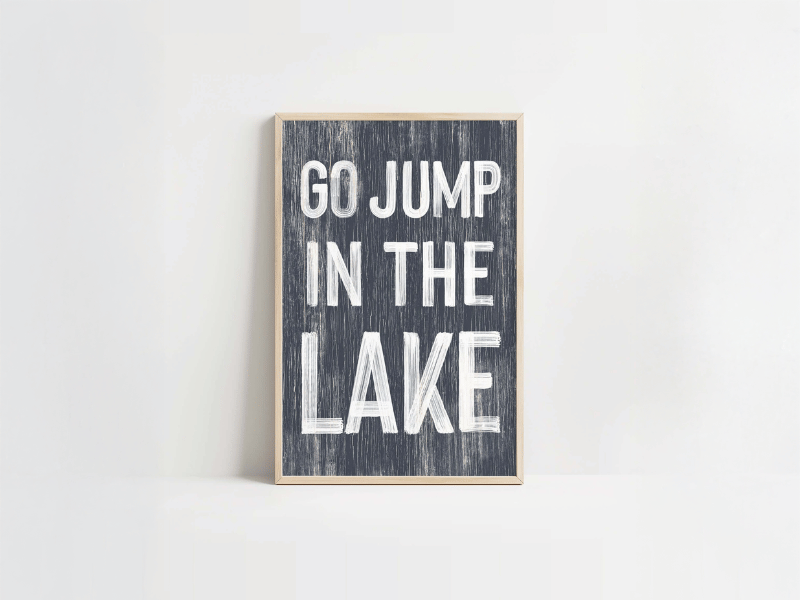 A framed sign with white text reads "GO JUMP IN THE LAKE" on a dark, weathered background.