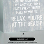 a sign with text that reads "KEEP THE SAND OUTSIDE HANG UP YOUR TOWEL HAVE ANOTHER DRINK ENJOY EVERY SUNSET MAKE MEMORIES RELAX, YOU'RE AT THE BEACH!" The sign is made of aluminum print and is described as waterproof and perfect for outdoor use.