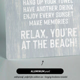 a sign with text that reads "KEEP THE SAND OUTSIDE HANG UP YOUR TOWEL HAVE ANOTHER DRINK ENJOY EVERY SUNSET MAKE MEMORIES RELAX, YOU'RE AT THE BEACH!" The sign is made of aluminum print and is described as waterproof and perfect for outdoor use.