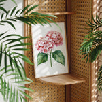 a framed painting of a pink hydrangea flower on a wooden shelf, surrounded by lush green leaves.