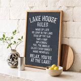A chalkboard sign with rules for a lake house, accompanied by a potted plant, a metal container, and a bowl of lemons, all set against a white brick wall.