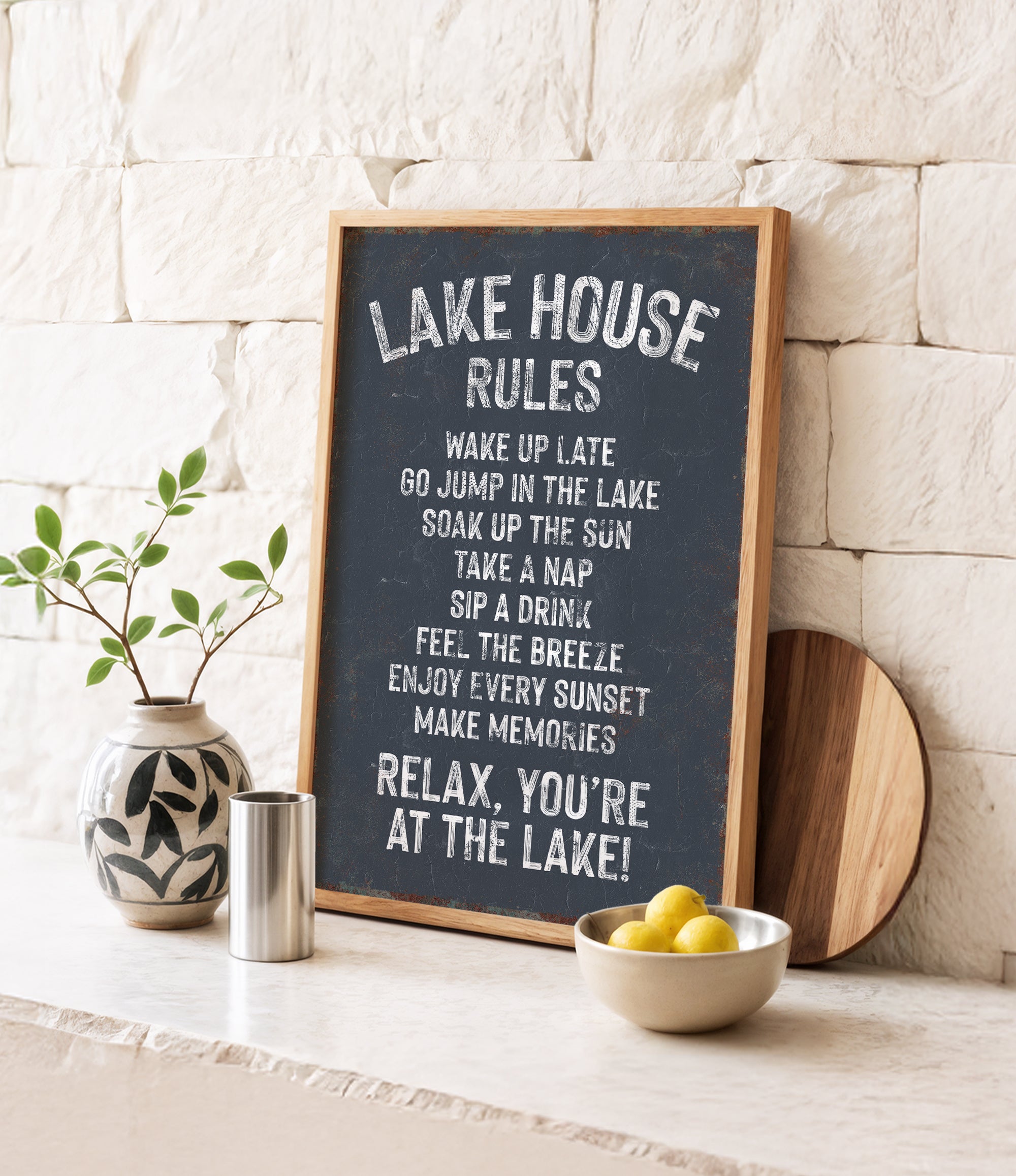A chalkboard sign with rules for a lake house, accompanied by a potted plant, a metal container, and a bowl of lemons, all set against a white brick wall.
