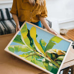 A woman is holding a framed painting of a vibrant, colorful tropical plant, likely a banana, in a room with a couch and other furniture.