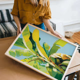 A woman is holding a framed painting of a vibrant, colorful tropical plant, likely a banana, in a room with a couch and other furniture.