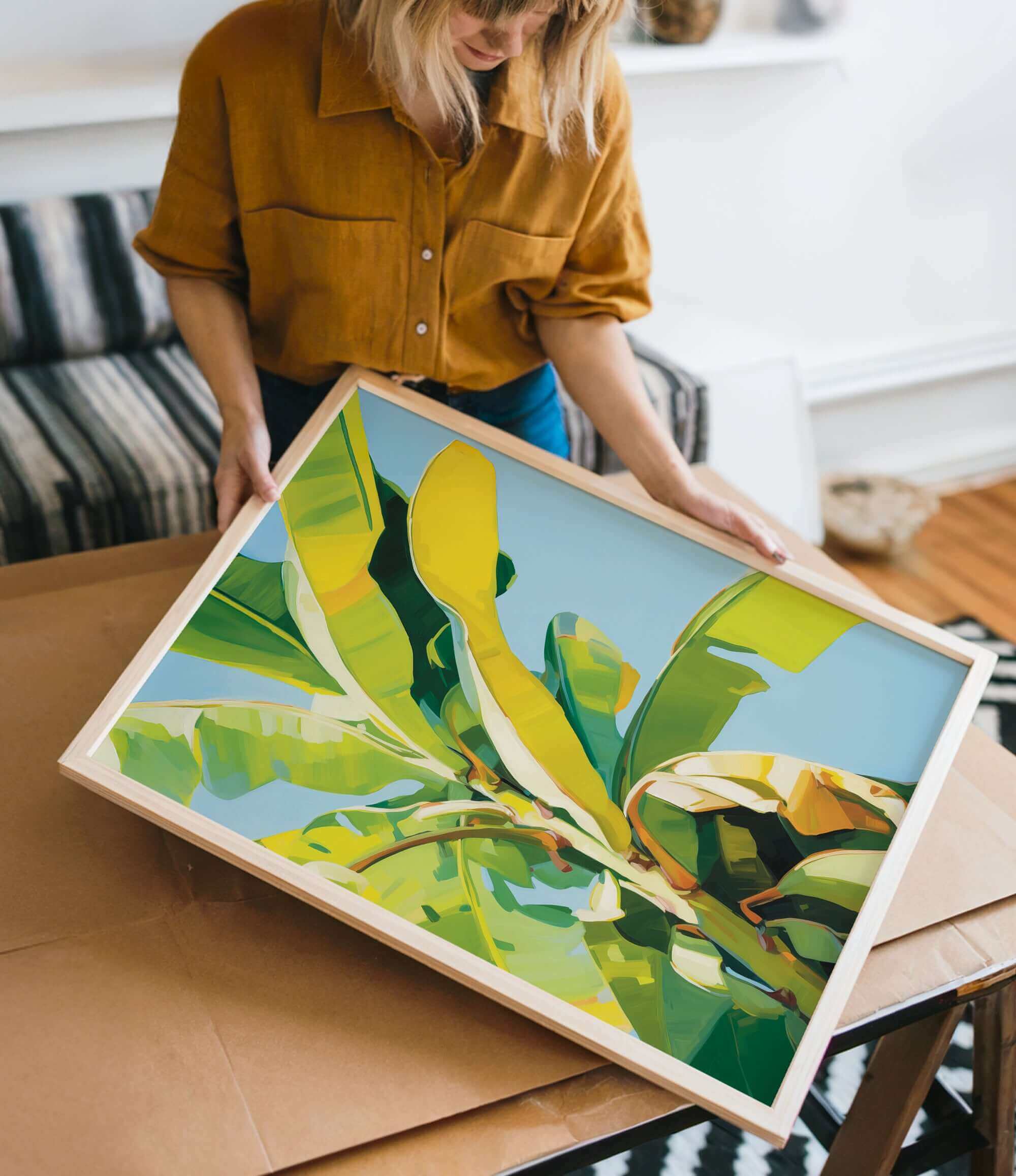 A woman is holding a framed painting of a vibrant, colorful tropical plant, likely a banana, in a room with a couch and other furniture.