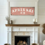 a cozy living room with a fireplace, a wooden mantel, and a framed sign that reads "APRÈS SKI". The mantel has a vase and a candle on it, adding to the warm and inviting atmosphere of the room.