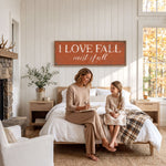 A woman and a young girl are sitting on a bed in a cozy bedroom, with a large orange sign above them that says "I LOVE FALL most of all".