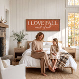 A woman and a young girl are sitting on a bed in a cozy bedroom, with a large orange sign above them that says "I LOVE FALL most of all".