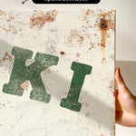 a close-up view of a white canvas print with a green and black text design. The text appears to be a stylized representation of the letters "K" and "I".