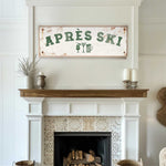 a cozy living room with a white fireplace adorned with a decorative tile pattern. Above the fireplace, there is a rustic sign that reads "APRES SKI", suggesting a connection to skiing or a ski lodge. The room also includes a wooden shelf with a vase and a potted plant, adding to the warm and inviting atmosphere.