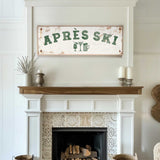 a cozy living room with a white fireplace adorned with a decorative tile pattern. Above the fireplace, there is a rustic sign that reads "APRES SKI", suggesting a connection to skiing or a ski lodge. The room also includes a wooden shelf with a vase and a potted plant, adding to the warm and inviting atmosphere.