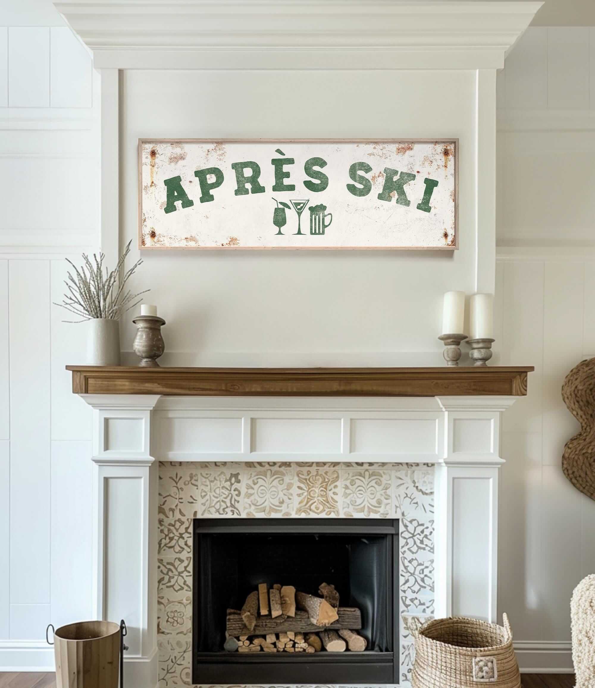 a cozy living room with a white fireplace adorned with a decorative tile pattern. Above the fireplace, there is a rustic sign that reads "APRES SKI", suggesting a connection to skiing or a ski lodge. The room also includes a wooden shelf with a vase and a potted plant, adding to the warm and inviting atmosphere.