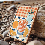 A framed illustration of a glass of iced tea with orange slices and a green leaf, set against a checkered background, is displayed on a rocky beach.