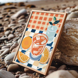 A framed illustration of a glass of iced tea with orange slices and a green leaf, set against a checkered background, is displayed on a rocky beach.