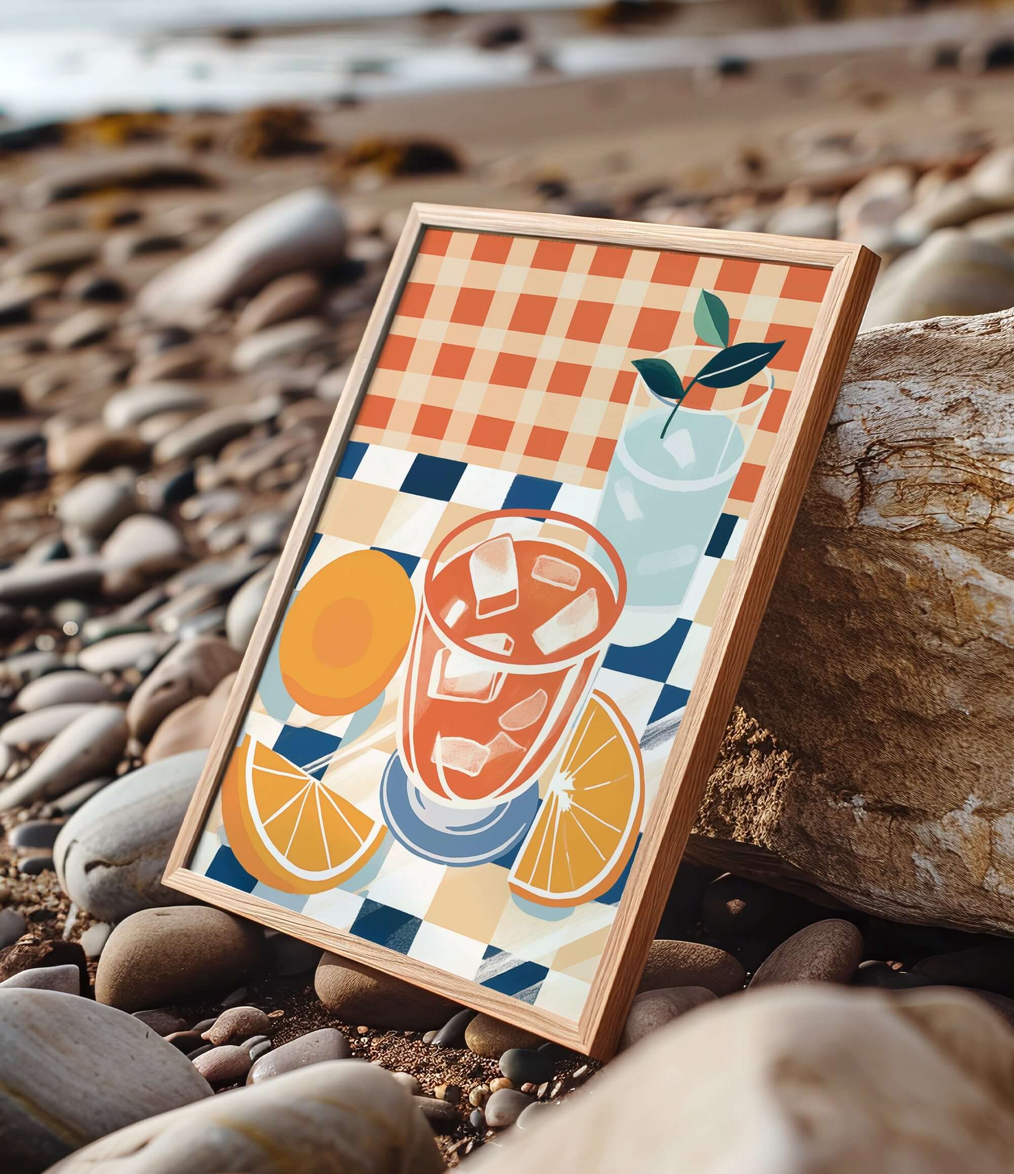 A framed illustration of a glass of iced tea with orange slices and a green leaf, set against a checkered background, is displayed on a rocky beach.