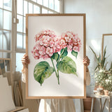 A person is holding up a framed painting of pink hydrangea flowers in a room with various potted plants and art supplies.