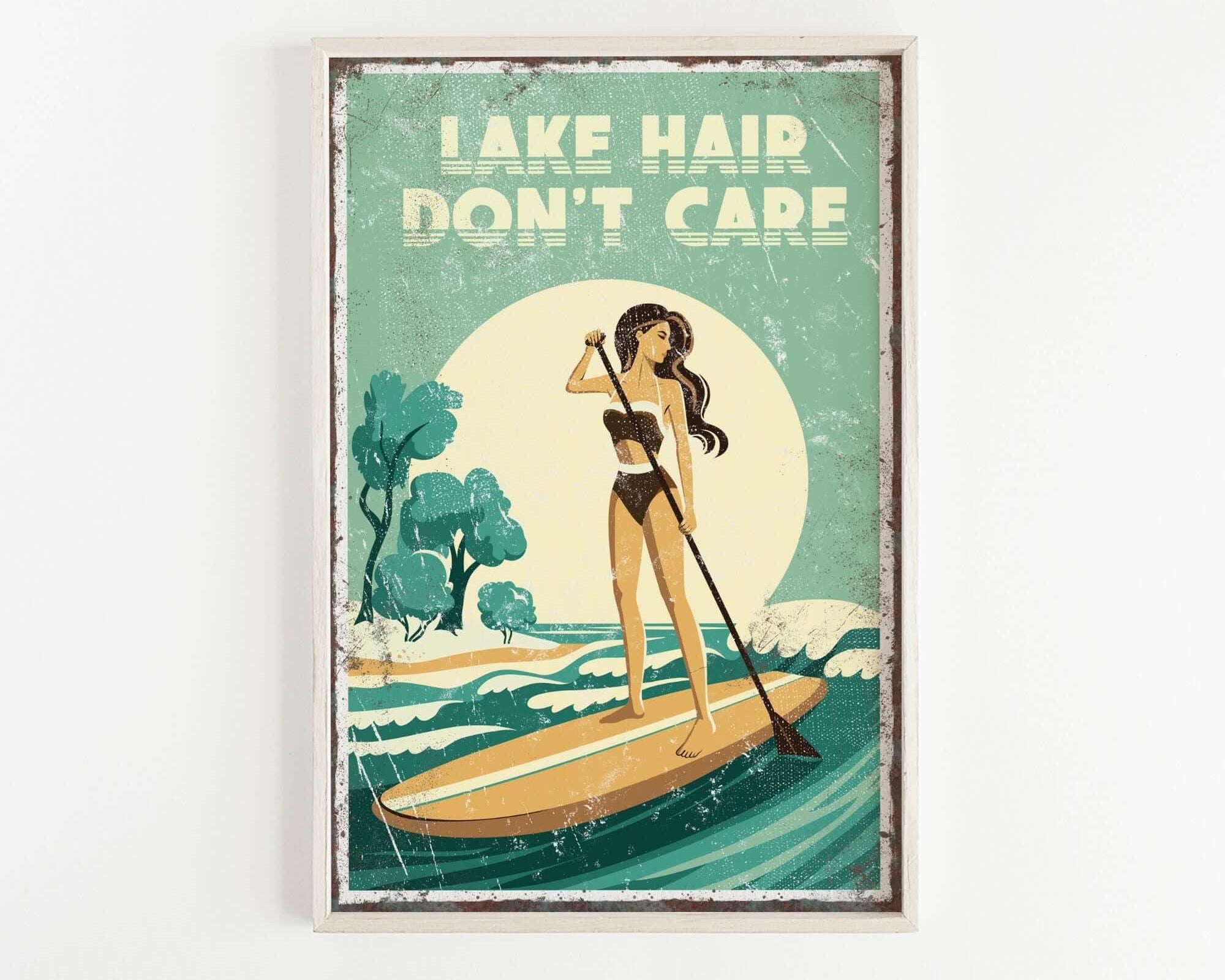 The image is a framed poster with a vintage-style illustration of a woman paddleboarding on a lake. The poster features the text "LAKESHAIR DON'T CARE" prominently displayed.