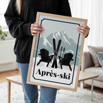 A person is holding a framed poster with a skiing scene featuring two black Adirondack chairs and skis, with a mountain backdrop.