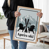 A person is holding a framed poster with a skiing scene featuring two black Adirondack chairs and skis, with a mountain backdrop.