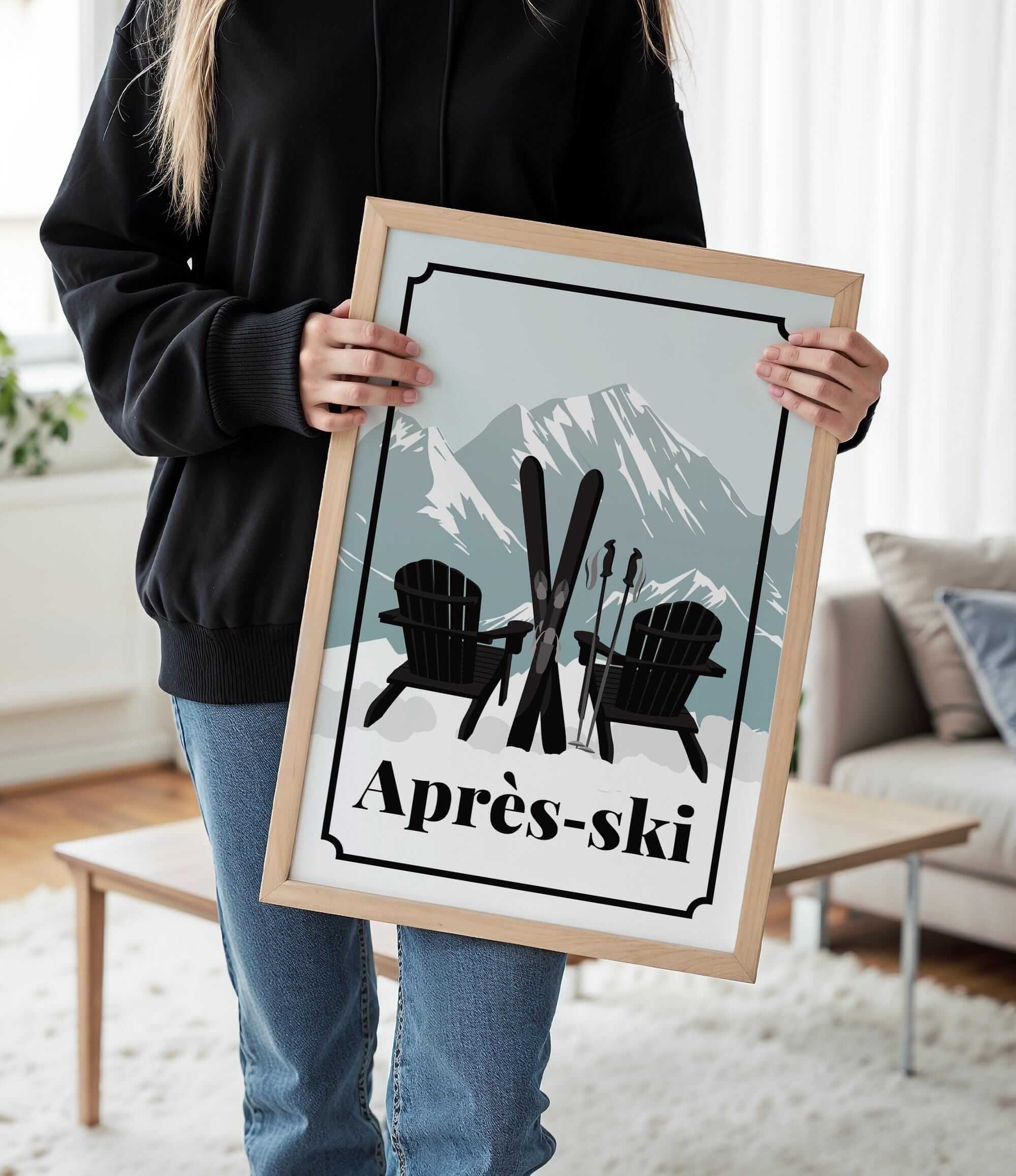 A person is holding a framed poster with a skiing scene featuring two black Adirondack chairs and skis, with a mountain backdrop.