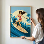A woman is standing in front of a framed illustration of a woman surfing on a surfboard, with palm trees and a sunset in the background.