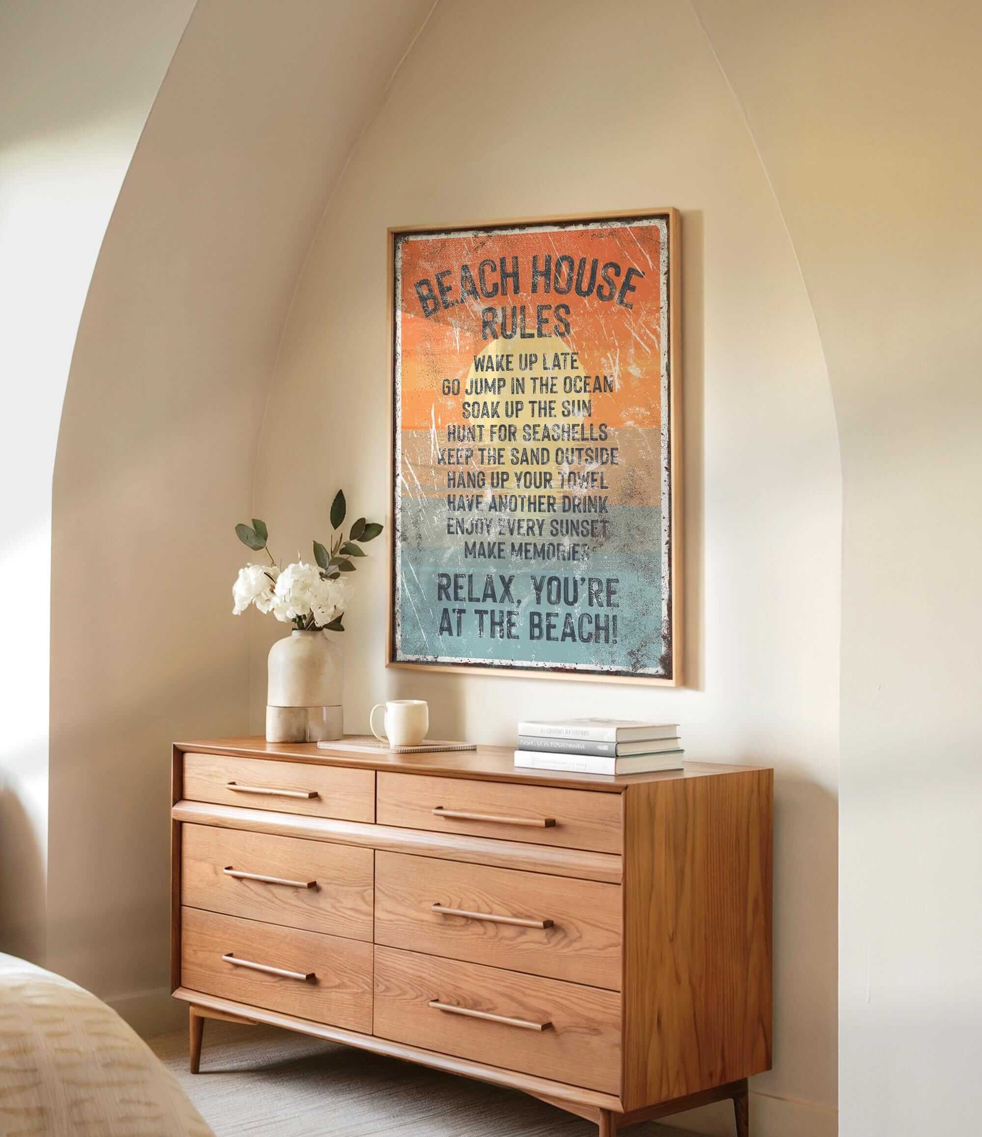 a wooden dresser with a framed sign that reads "BEACH HOUSE RULES" hanging on the wall above it. The dresser has a vase of white flowers and a stack of books on top.