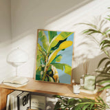 a wooden shelf with a framed painting of a banana tree, a lamp, and various books and plants.