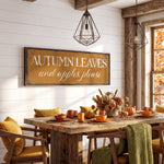 a rustic dining room with a wooden table set for autumn-themed dinner. The table is adorned with orange and yellow fall-themed dishes, and a large sign above the table reads "Autumn Leaves and Apples Please". The room is decorated with pendant lights, a wooden beam ceiling, and a window with autumn trees visible outside.