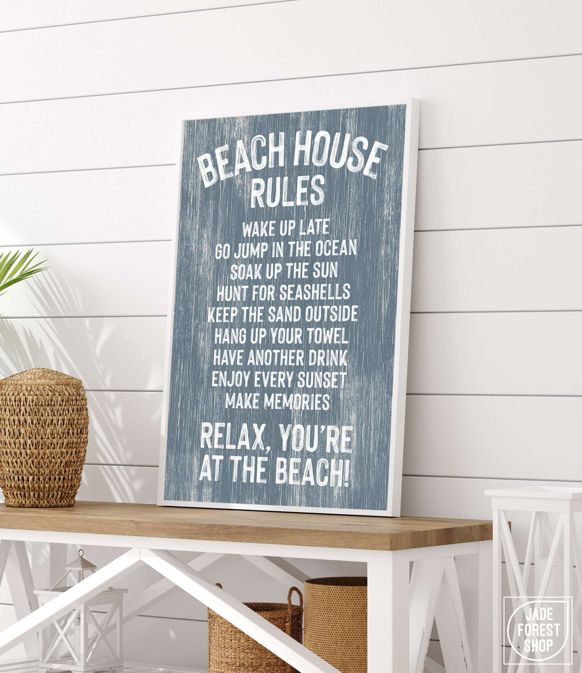 The Beach House Rules Sign, Painted Wood in Harbor Blue, sits on a white table amid coastal decor and plants—a perfect touch for your beach house decor or as eye-catching coastal wall art.