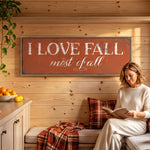 A woman is sitting on a bench, reading a book, with a rustic wooden wall behind her. The wall has a large sign that says "I LOVE FALL most of all".