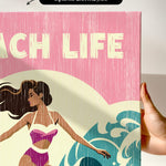 a hand holding a canvas print with a beach scene and a woman in a swimsuit.