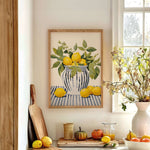 a kitchen counter with a framed painting of lemons and a vase of flowers, a cutting board with a knife, a bowl of fruit, and a potted plant.
