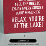 The image is a red sign with white text that reads "Sip a drink, feel the breeze, enjoy every sunset, make memories, relax, you're at the lake!"