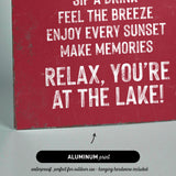 The image is a red sign with white text that reads "Sip a drink, feel the breeze, enjoy every sunset, make memories, relax, you're at the lake!"