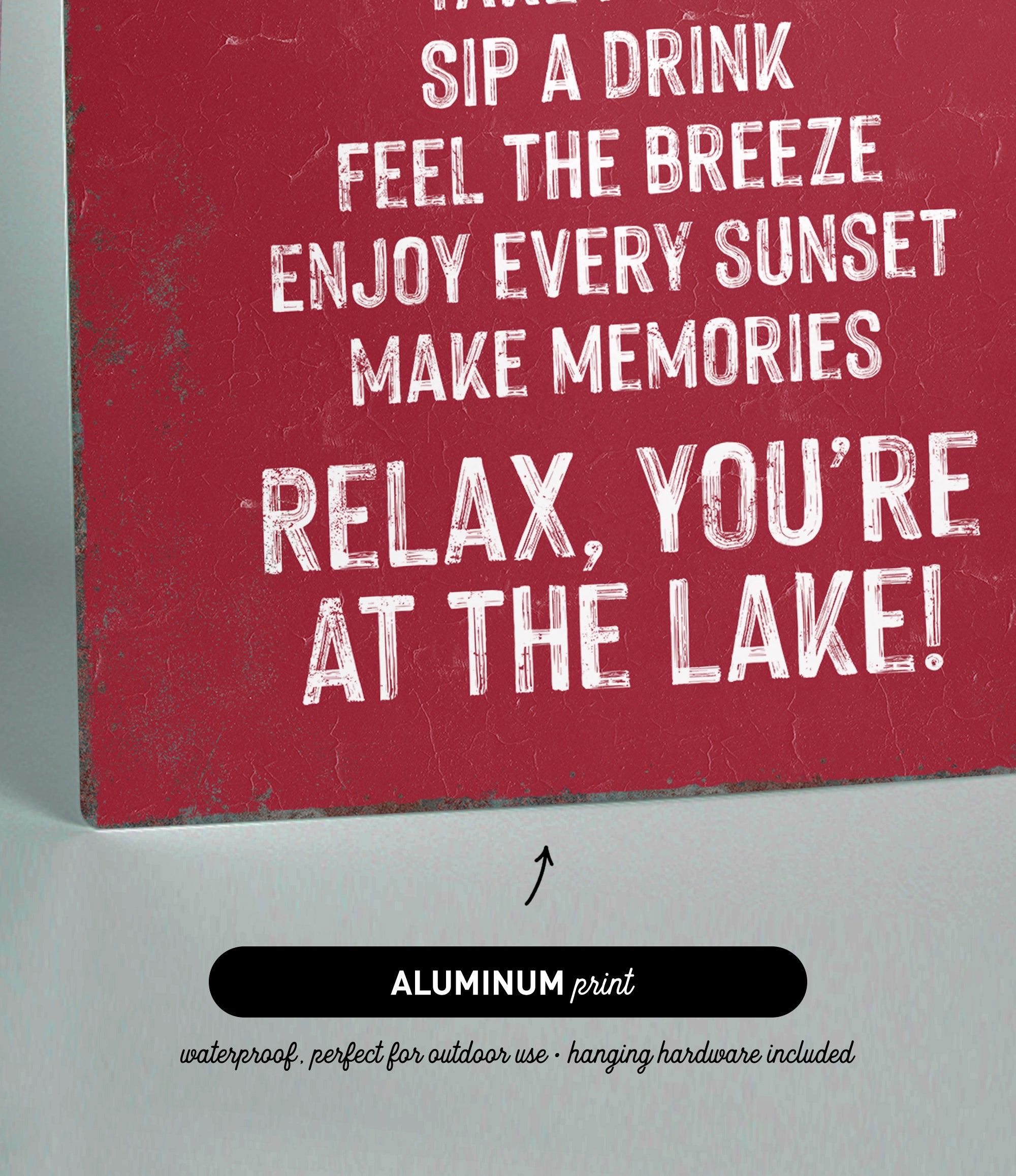 The image is a red sign with white text that reads "Sip a drink, feel the breeze, enjoy every sunset, make memories, relax, you're at the lake!"