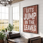 a rustic-style room with a large wooden sign that reads "Let's JUMP IN THE LEAVES" in bold white letters. The sign is mounted on a white wall above a leather couch, which is positioned in front of a window that offers a view of a field. The room also contains several potted plants, adding a touch of greenery to the space.