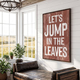 a rustic-style room with a large wooden sign that reads "Let's JUMP IN THE LEAVES" in bold white letters. The sign is mounted on a white wall above a leather couch, which is positioned in front of a window that offers a view of a field. The room also contains several potted plants, adding a touch of greenery to the space.