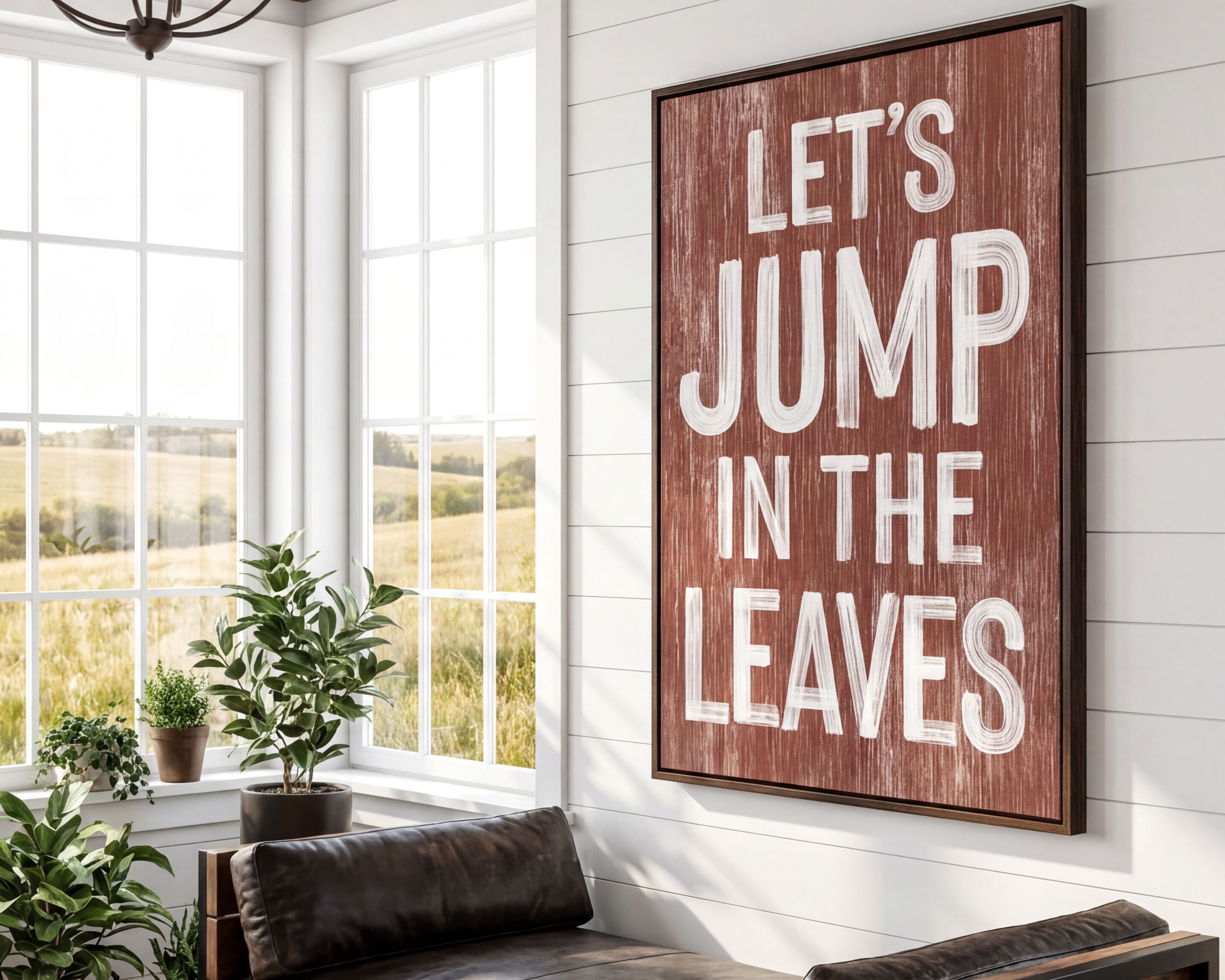 a rustic-style room with a large wooden sign that reads "Let's JUMP IN THE LEAVES" in bold white letters. The sign is mounted on a white wall above a leather couch, which is positioned in front of a window that offers a view of a field. The room also contains several potted plants, adding a touch of greenery to the space.