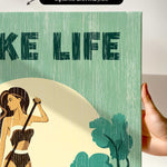 a hand holding a framed print with a green and yellow color scheme. The print has a textured appearance and includes a woman holding a cane.