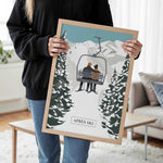 A person is holding a framed picture of a ski lift with two people on it, in a snowy mountain setting.