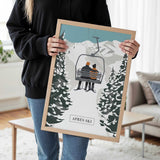 A person is holding a framed picture of a ski lift with two people on it, in a snowy mountain setting.