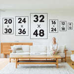 A woman sits on a sofa under the Après Ski Lift Couple Wall Art, Yellow Print, which is displayed in various frame sizes in a modern living room.