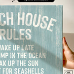 A hand holds the Beach House Rules Sign in Tide, crafted from painted wood—coastal wall art perfect for your space.