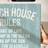 A hand holds the Beach House Rules Sign in Tide, crafted from painted wood—coastal wall art perfect for your space.