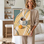 A woman is holding a framed print of a still life composition featuring a cup, lemon, and orange on a striped tablecloth.