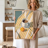A woman is holding a framed print of a still life composition featuring a cup, lemon, and orange on a striped tablecloth.