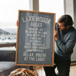A man holding a sign that says "LAKESHOUSE RULES" in front of a window overlooking a lake with boats.