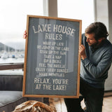 A man holding a sign that says "LAKESHOUSE RULES" in front of a window overlooking a lake with boats.