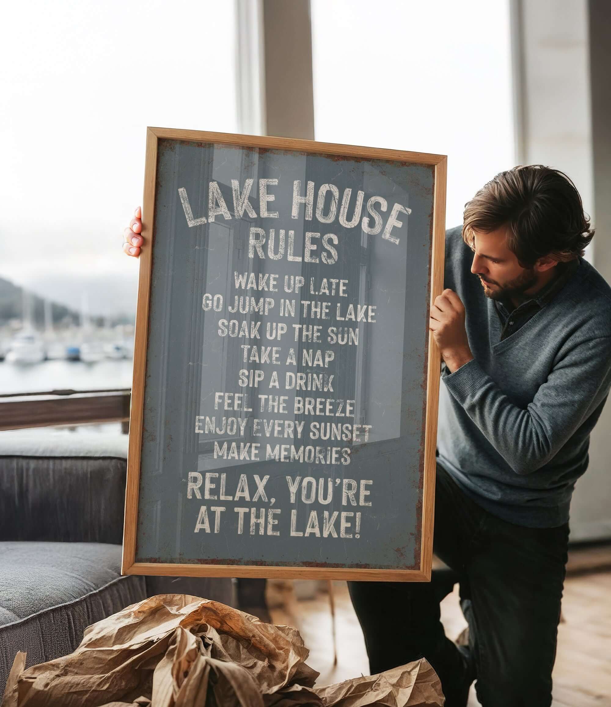 A man holding a sign that says "LAKESHOUSE RULES" in front of a window overlooking a lake with boats.